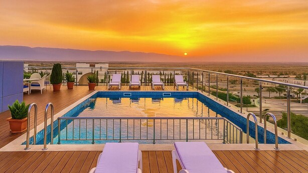 Una piscina en la azotea con sillones de descanso y plantas en macetas, con vistas a un paisaje urbano al atardecer.