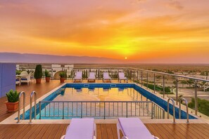 Una piscina en la azotea con sillones de descanso y plantas en macetas, con vistas a un paisaje urbano al atardecer.