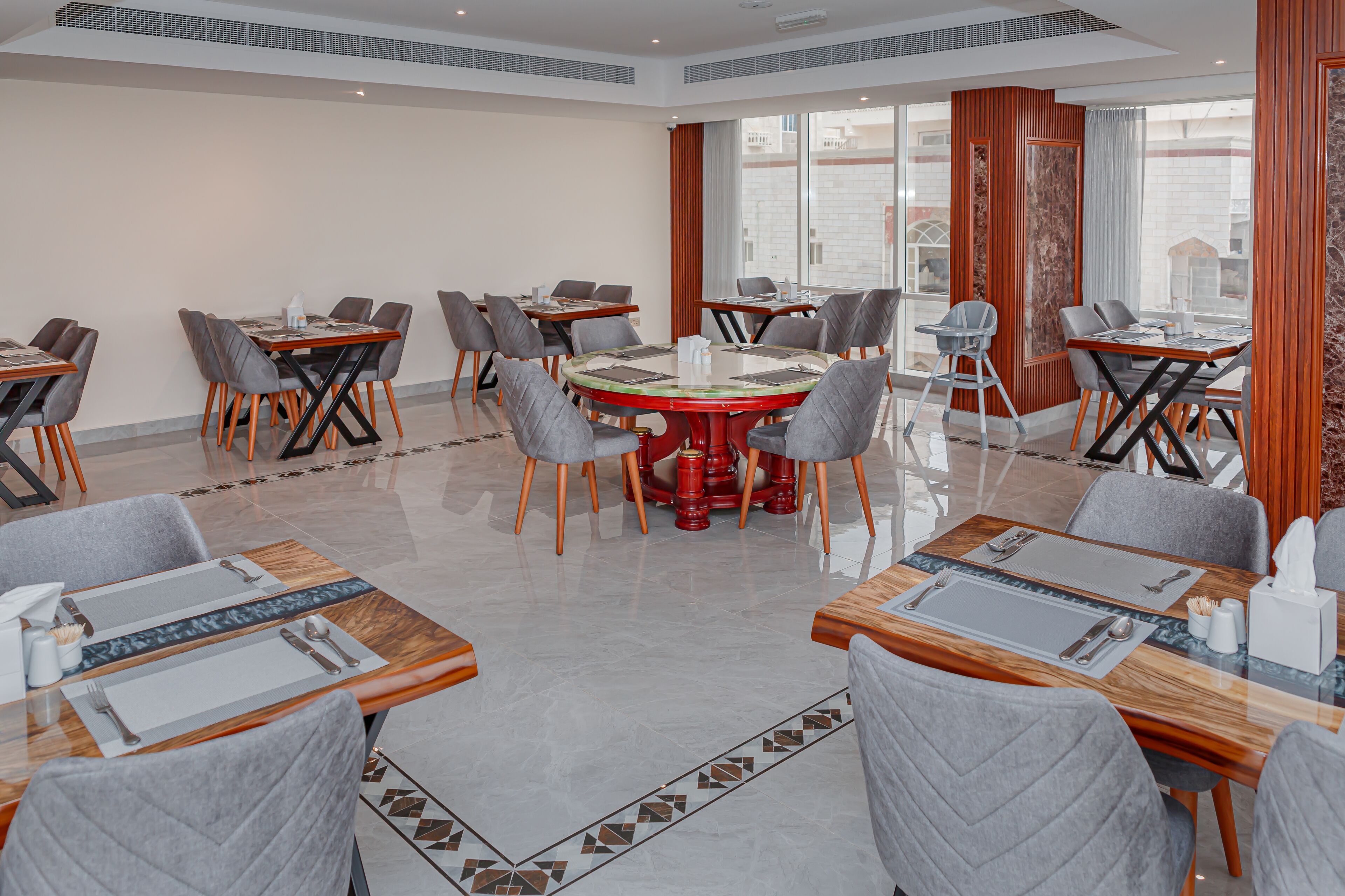 A hotel dining area with multiple tables and chairs, large windows, and a modern interior design.