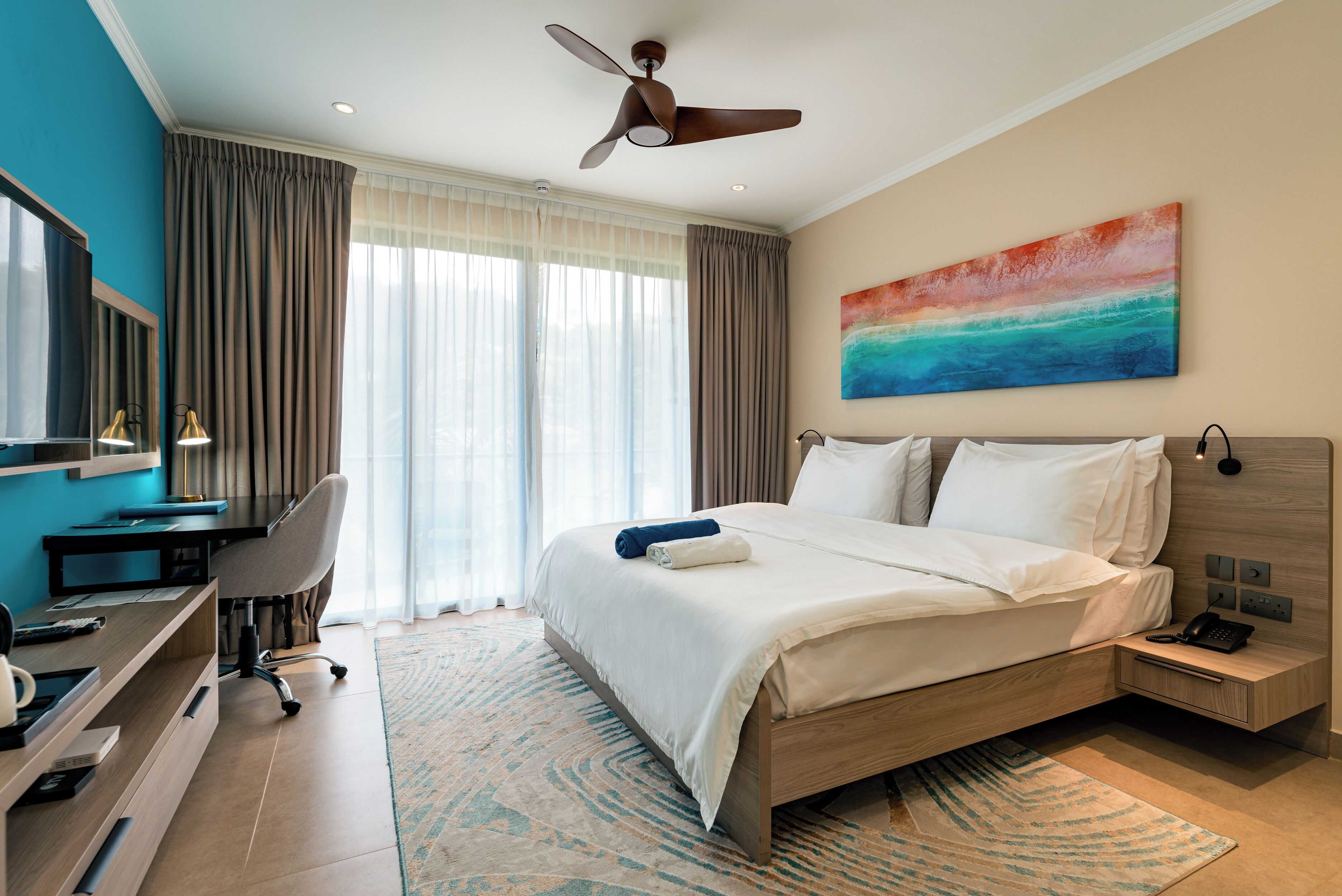 A modern hotel room with a bed, desk, and chair, a ceiling fan, and a large window with curtains.