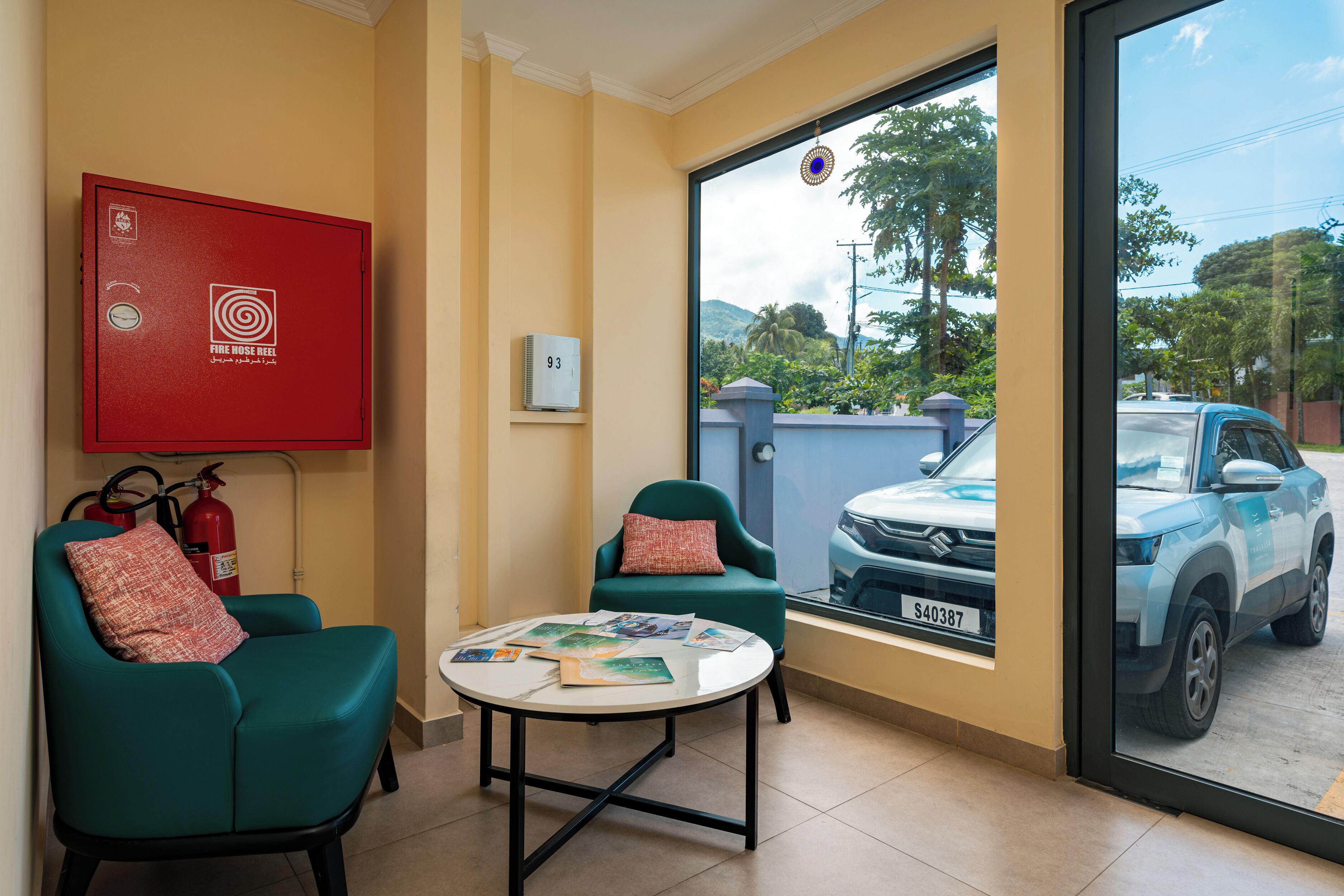 Une pièce avec des chaises de couleur turquoise, une table ronde avec des magazines, un coffre rouge contenant un tuyau d’incendie, et une grande fenêtre donnant sur une voiture garée et de la verdure.