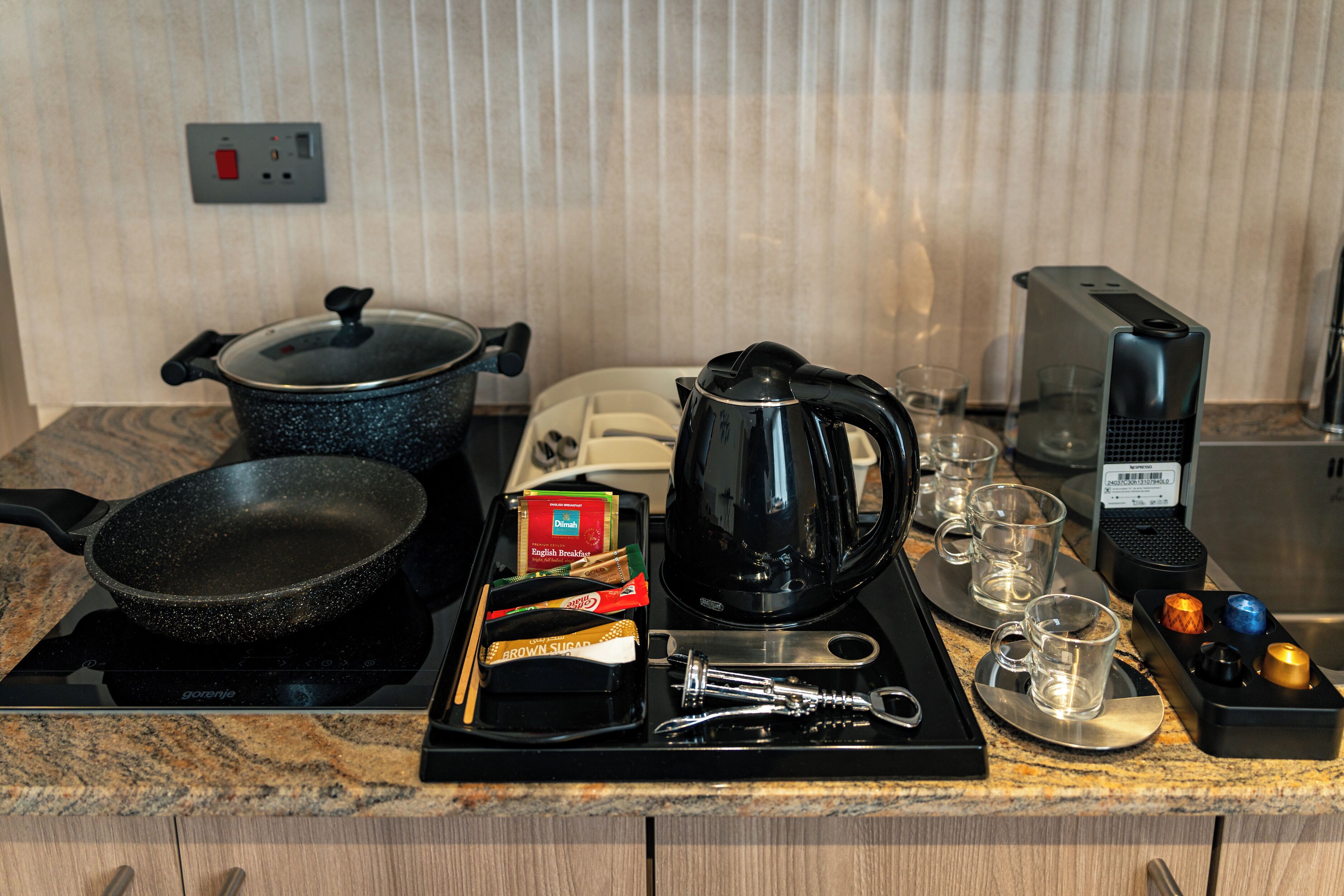 Een aanrecht met een koekenpan, een pan, een koffiezetapparaat, een theedoos, een waterkoker, een broodrooster en een set glazen.