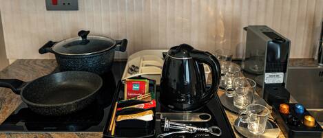 A kitchen counter with a frying pan, a pot, a coffee maker, a box of tea, a kettle, a toaster, and a set of glasses.