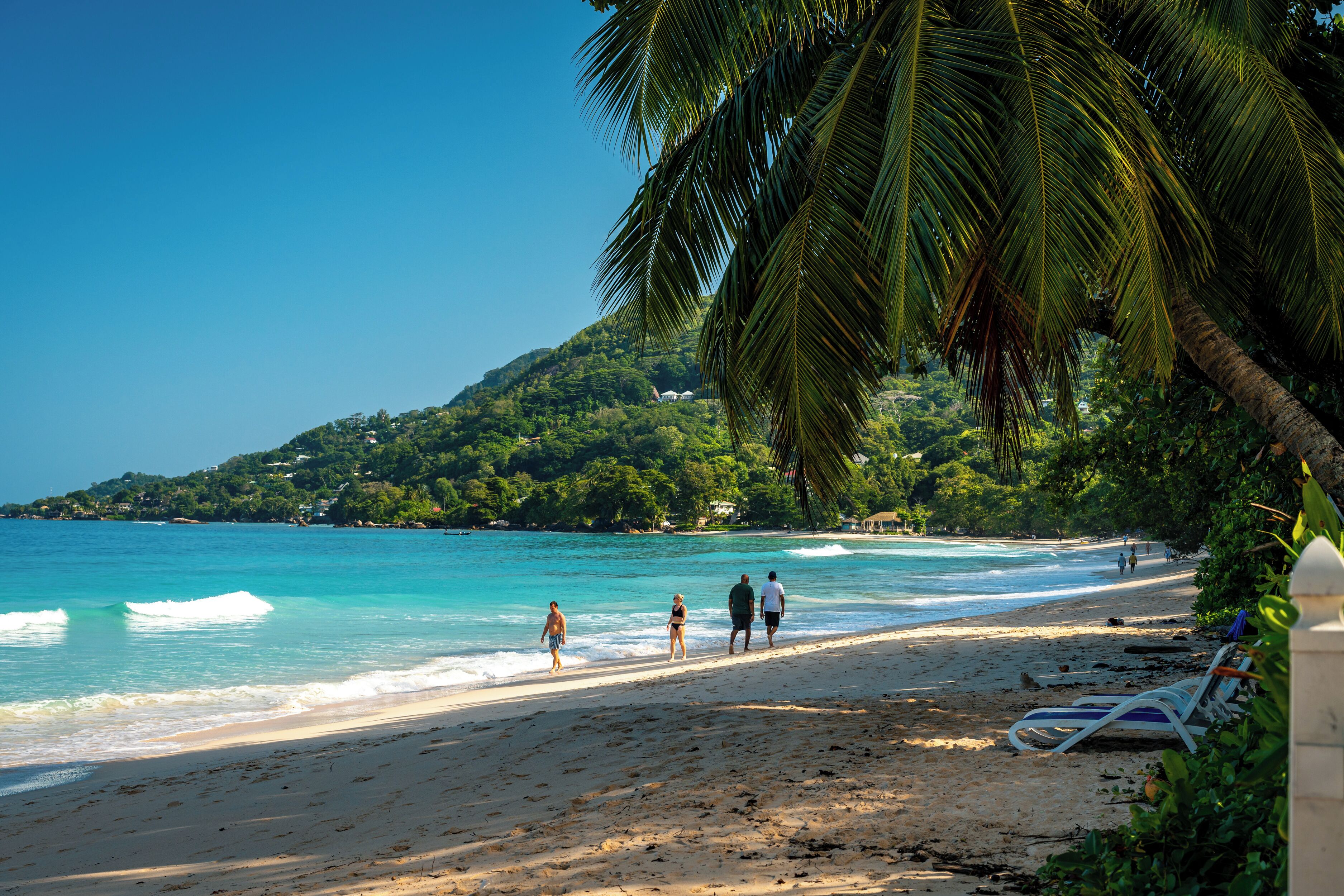 A beach with clear turquoise water, a sandy shore, and a few people walking. There are palm trees and a hill with greenery in the background.