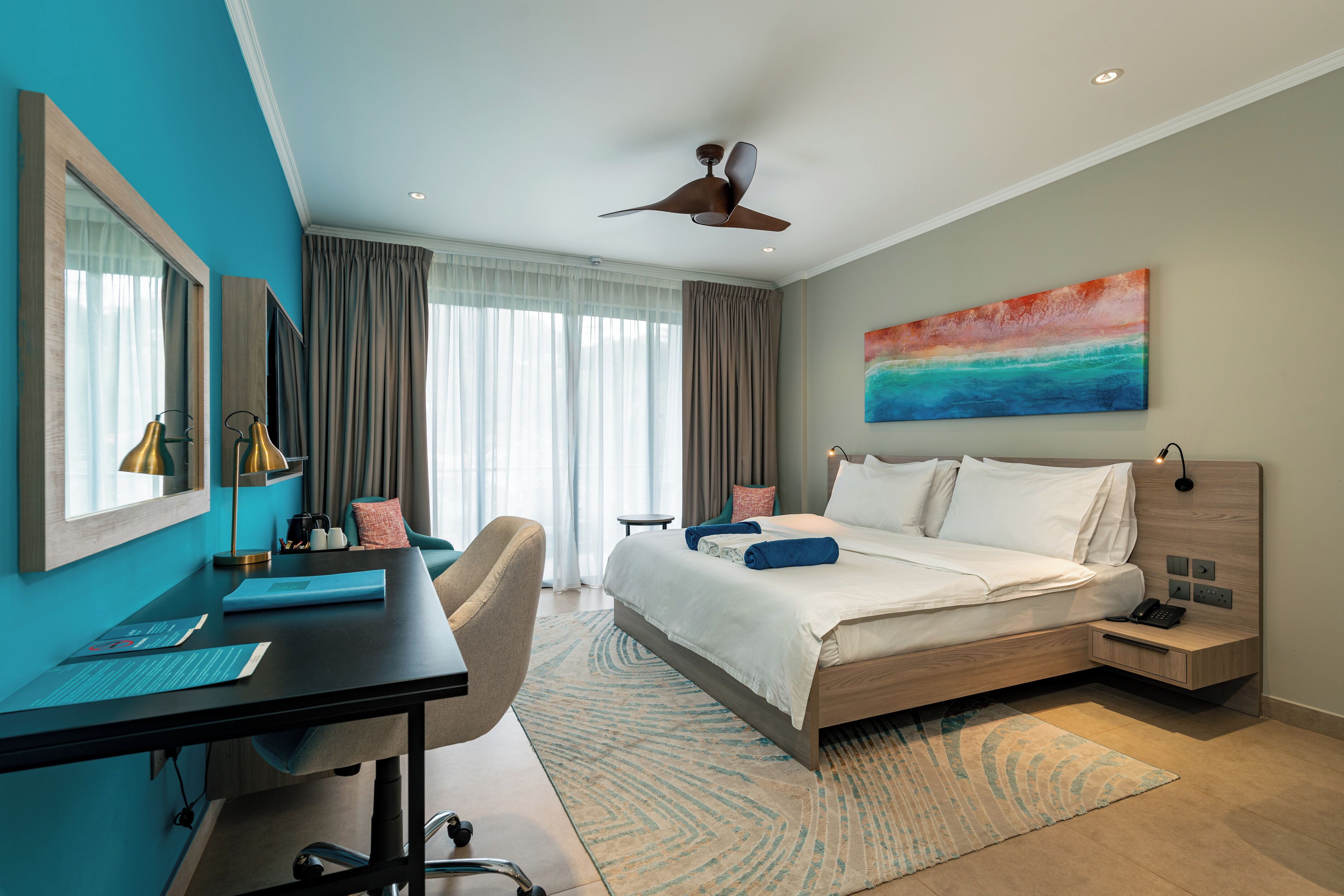 A modern hotel room with a large bed, a desk, a chair, a desk lamp, and a ceiling fan.