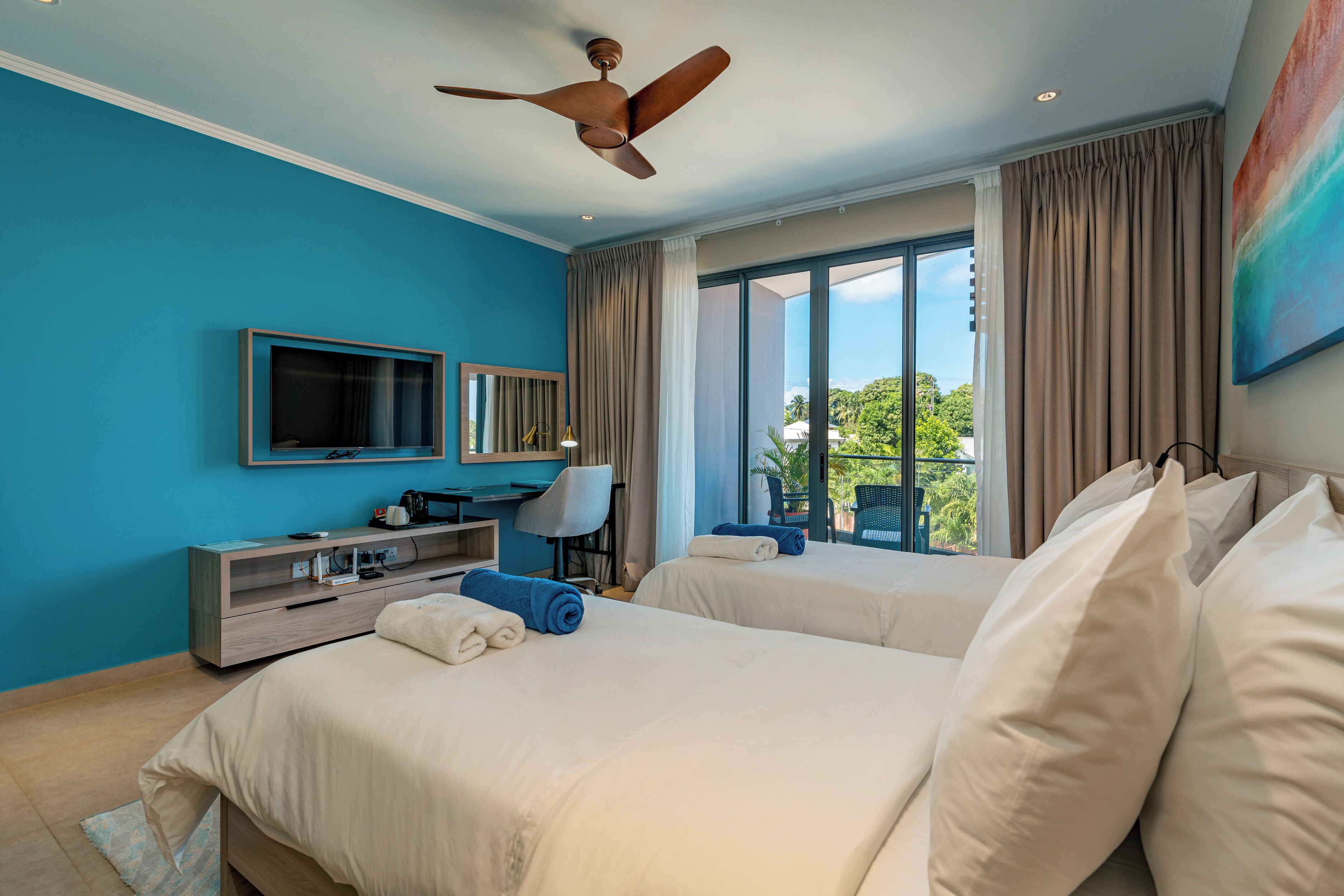 A hotel room with two beds, a ceiling fan, a TV, a desk, and a large window with curtains.