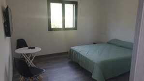 Um quarto pequeno e modesto, com uma cama de solteiro, uma cadeira, uma mesa pequena e uma janela.