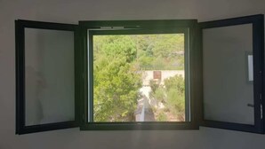 Quarto com uma grande janela que oferece vista para uma paisagem verde com árvores e um edifício.