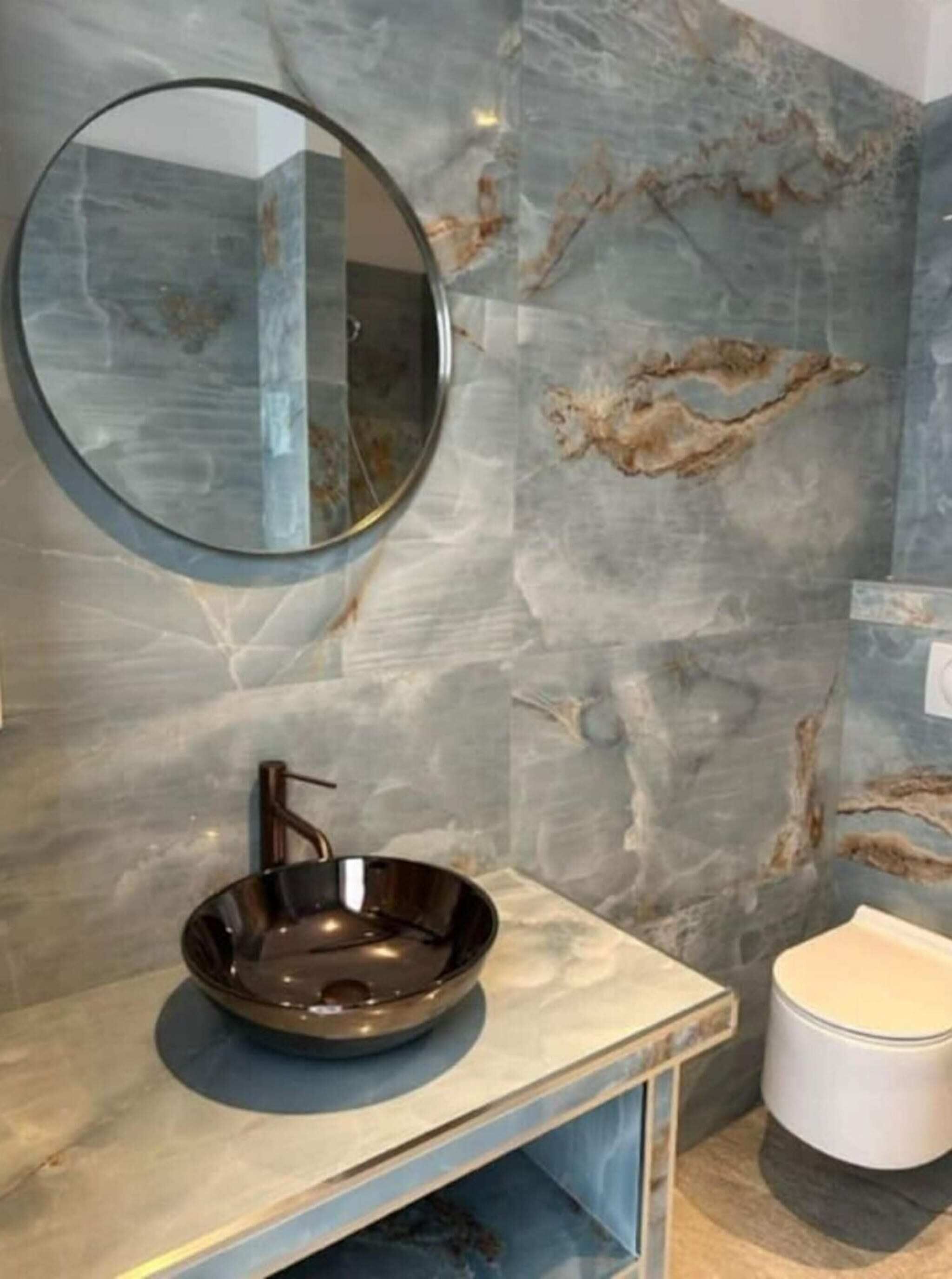 Une salle de bain avec un mur en marbre, un miroir rond, un lavabo moderne et des toilettes.