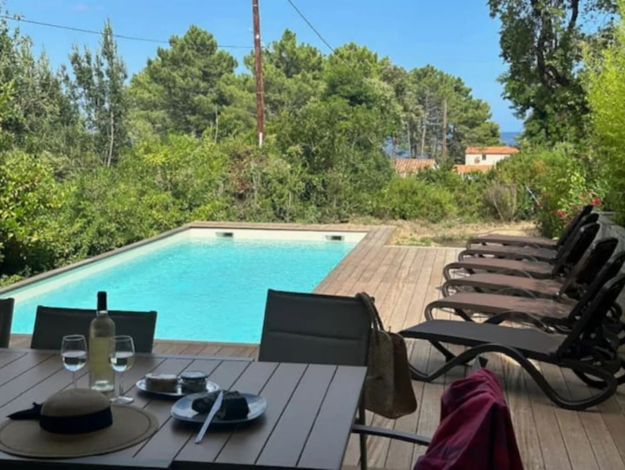 Un espace repas aménagé au bord de la piscine, avec une table dressée pour deux personnes, une bouteille de vin et un chapeau posé sur la table.