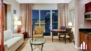 A hotel room with a sofa, armchairs, a glass coffee table, a dining table with chairs, a kitchen area, and a view of the cityscape through large windows. - MGM Signature-36-705 F1 Track & Strip View Balcony (Las Vegas)
