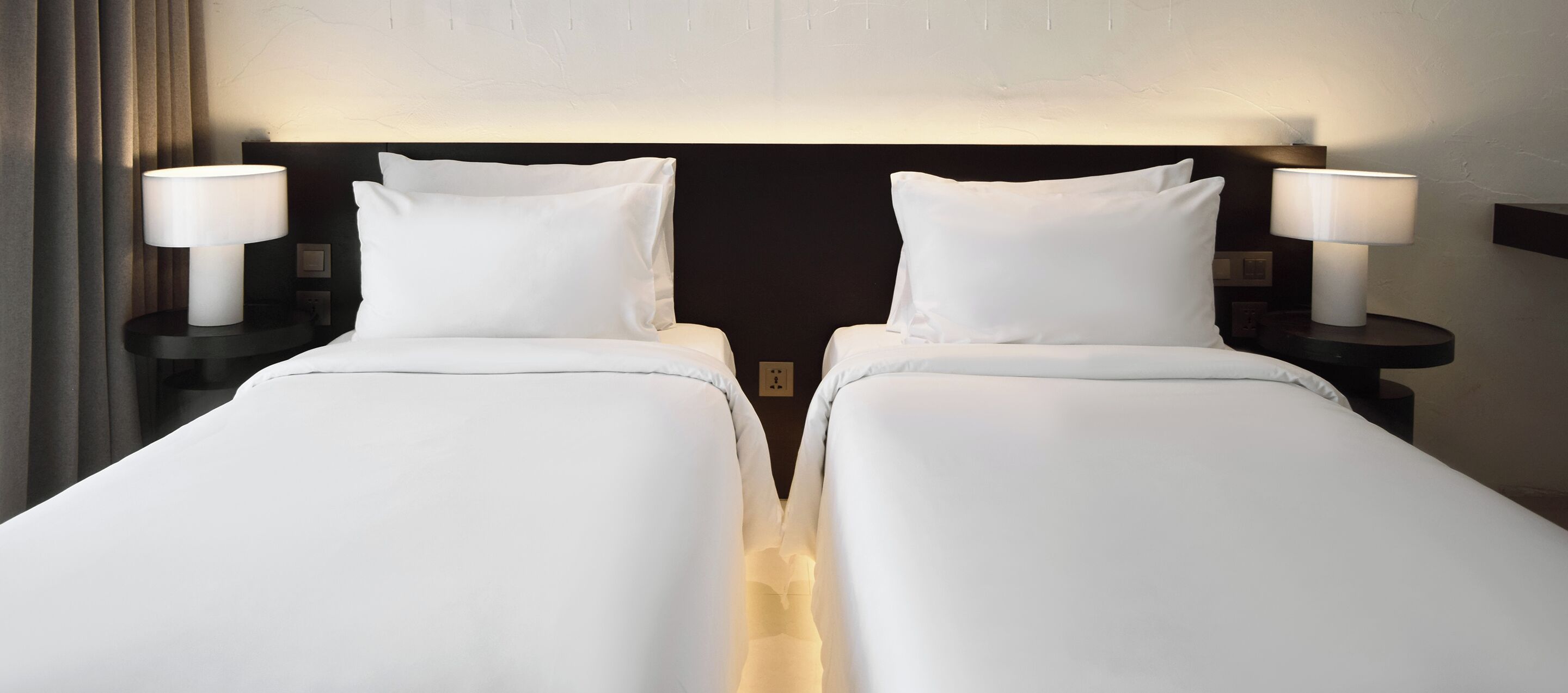 A hotel room with two single beds, each with a white bedspread and pillow, flanked by nightstands with lamps.