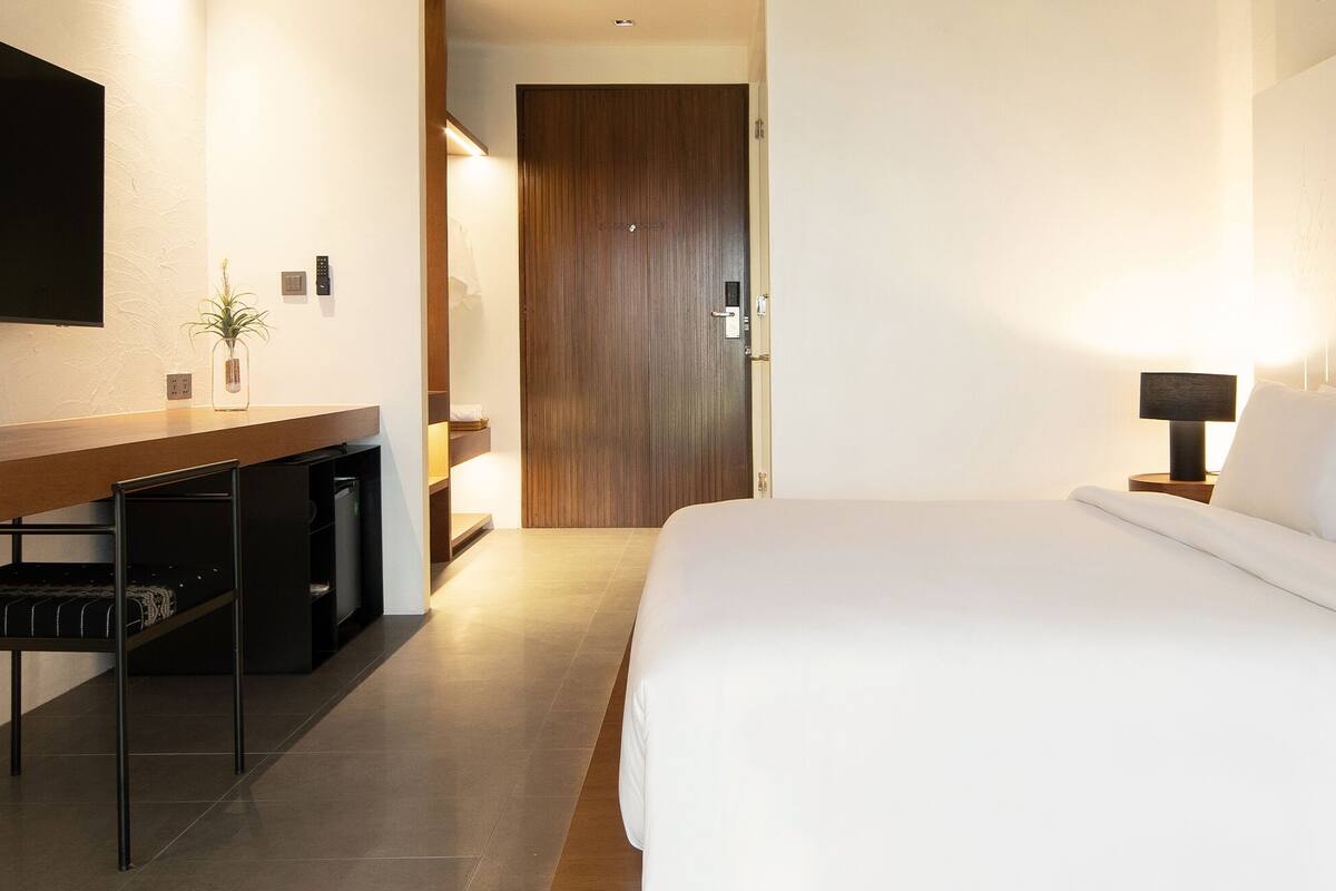 Habitación de hotel con una cama grande, un televisor montado en la pared, un escritorio con una planta y una mesita de noche con una lámpara.