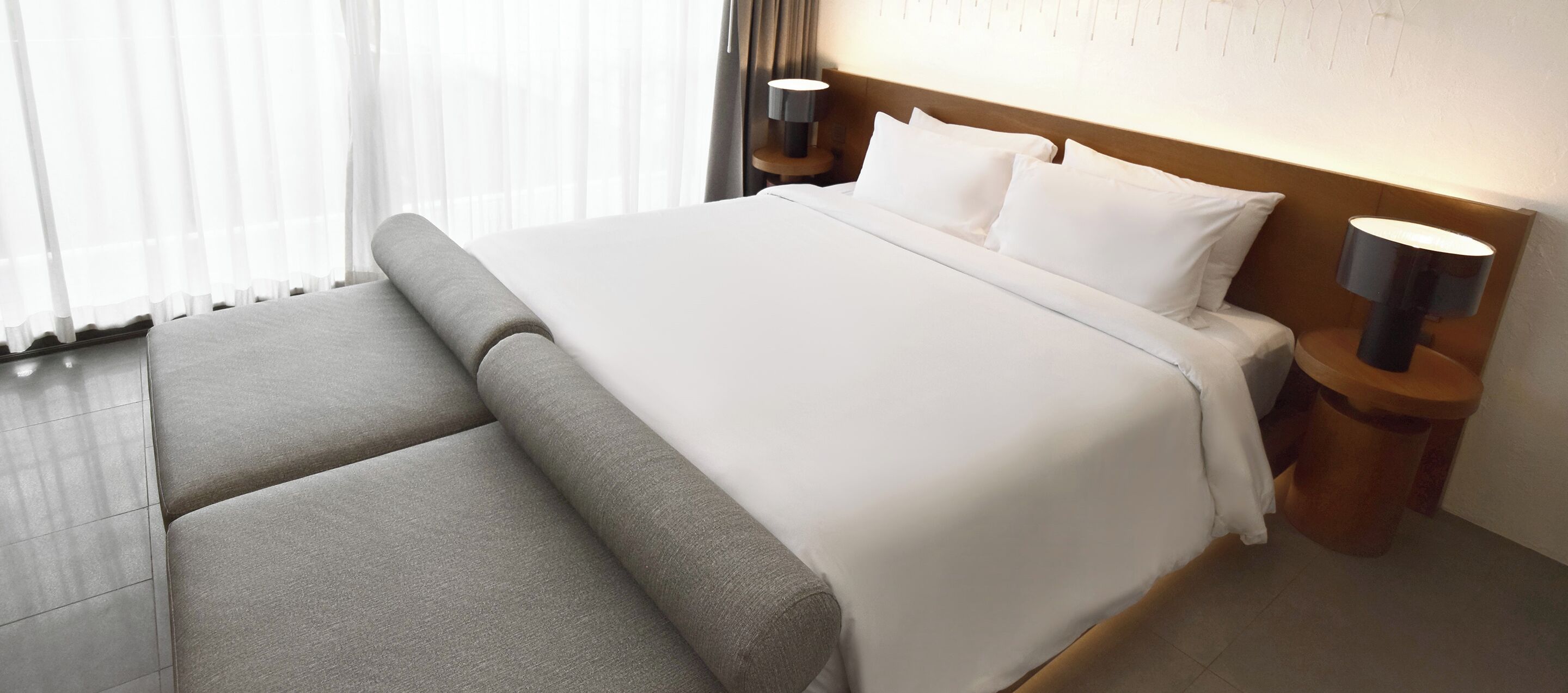 A hotel room with a large bed, a nightstand with a lamp, and a view of the outside through sheer curtains.