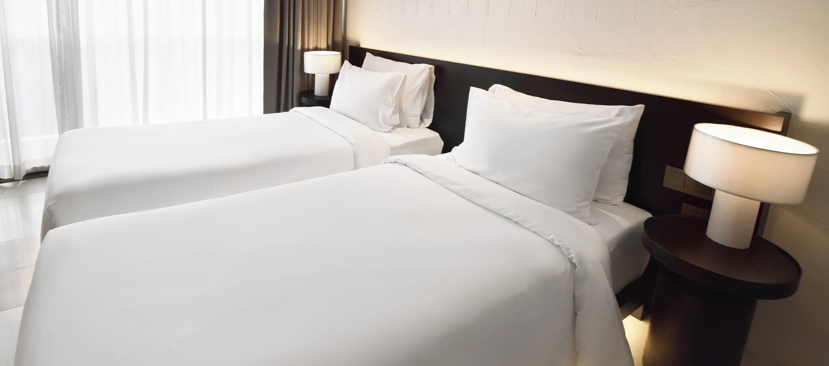 A hotel room with two beds, each with white linens and pillows, a nightstand with a lamp, and a window with curtains.
