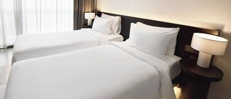 A hotel room with two beds, each with white linens and pillows, a nightstand with a lamp, and a window with curtains.