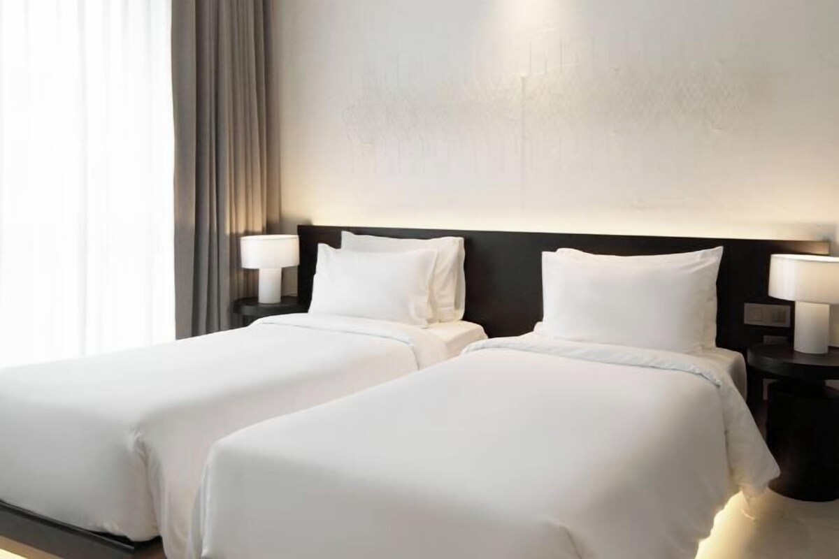 Una habitación de hotel con dos camas, cada una con ropa de cama blanca, cabeceros oscuros y lámparas de noche.