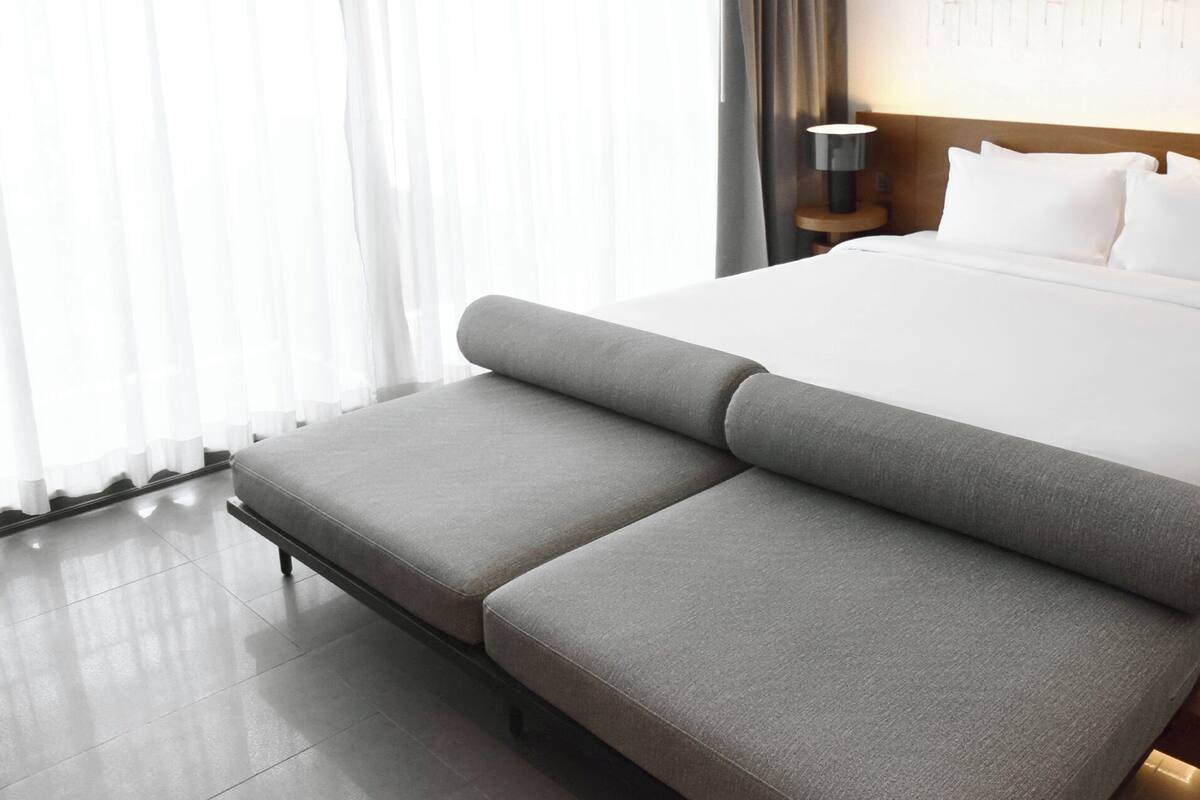 Un dormitorio moderno de hotel con una cama grande, dos mesitas de noche con lámparas, un banco al pie de la cama y cortinas translúcidas en la ventana.