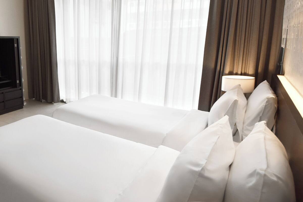 Habitación de hotel con cama, televisión, lámpara y vistas al exterior a través de cortinas translúcidas.
