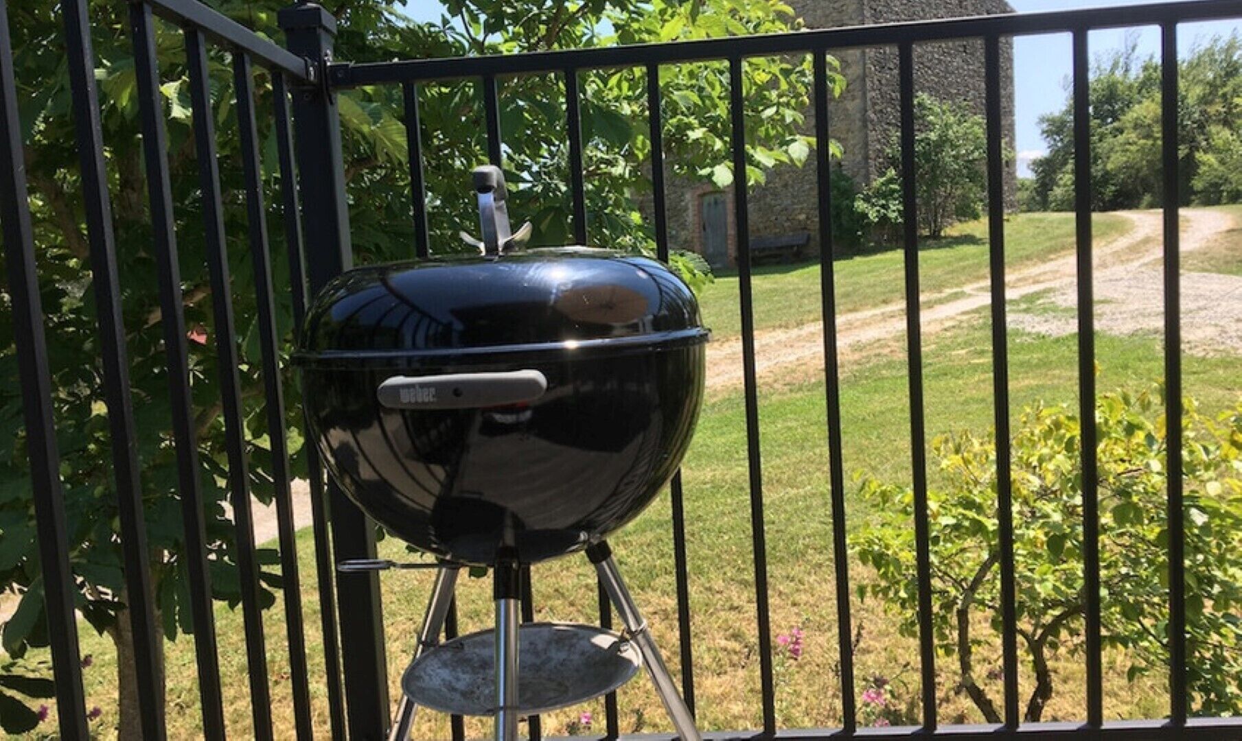 Una parrilla negra sobre un soporte, rodeada por una cerca metálica, con un área verde y árboles de fondo.
