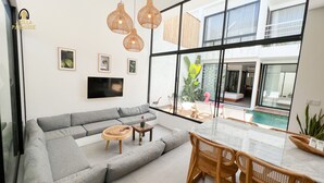 Uma sala de estar moderna com um sofá seccional cinza, uma TV fixada na parede, luminárias pendentes de vime e vista para uma piscina e varanda através de grandes janelas.