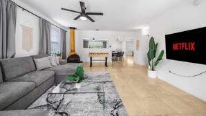 Ein modernes Wohnzimmer mit Ecksofa, Couchtisch, Kamin, Essbereich und einem großen Fernsehbildschirm, der 'NETFLIX' zeigt.