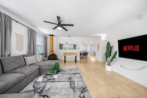 Una sala de estar moderna con un sofá modular, una mesa de centro, una chimenea, un comedor y una gran pantalla de televisión mostrando 'NETFLIX'.