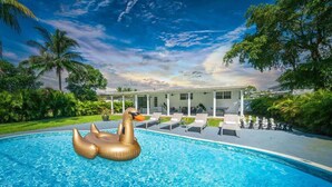 Ein Swimmingpool mit einem goldenen Schwimmswan, eine überdachte Terrasse mit Liegestühlen und ein Haus mit weißer Fassade.