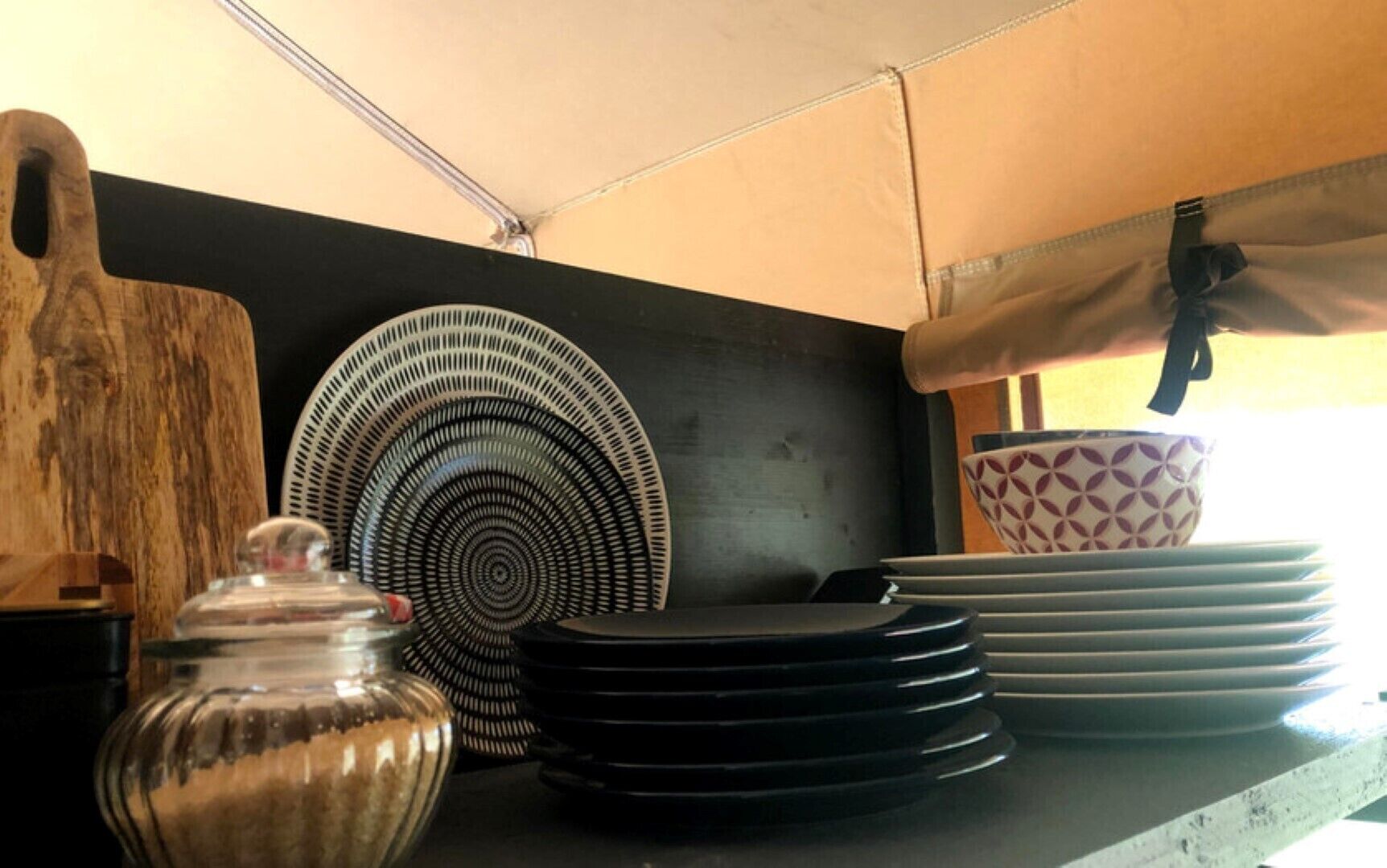 Una cocina con encimera negra, tabla de cortar de madera, tarro de vidrio, plato negro, cuenco estampado y una pila de platos.