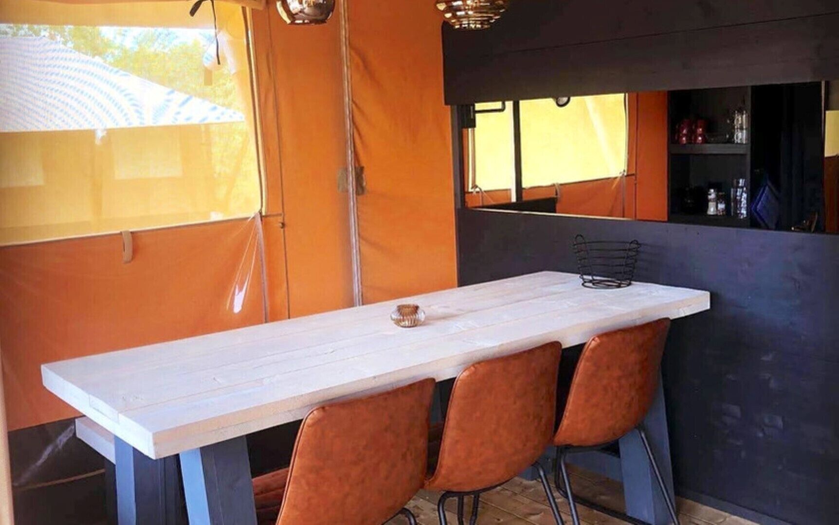 Un coin repas avec une table blanche et quatre chaises marron, un luminaire suspendu et une fenêtre donnant sur des arbres.