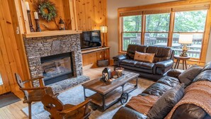 A cozy living room with a stone fireplace, leather sofas, and a large window offering a view of the outdoors. - Spring Brook Mountain Retreat + Hot Tub +Sauna+ Fire Pit (Mars Hill)