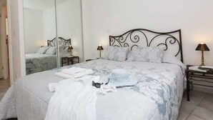 Una cama bien hecha con una colcha blanca y gris estampada, un plaid blanco y una almohada blanca. Hay dos mesitas de noche con lámparas, una a cada lado de la cama.