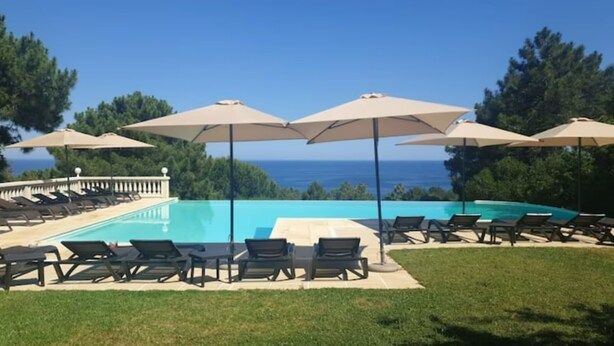 Zona junto a la piscina con sillones y sombrillas, con vistas a un cielo azul despejado y al océano.