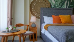 A neatly arranged bedroom with a bed, pillows, a bedside table with a lamp, a small table with a chair, and a decorative wall hanging. - Cove Griya Elite (Denpasar)