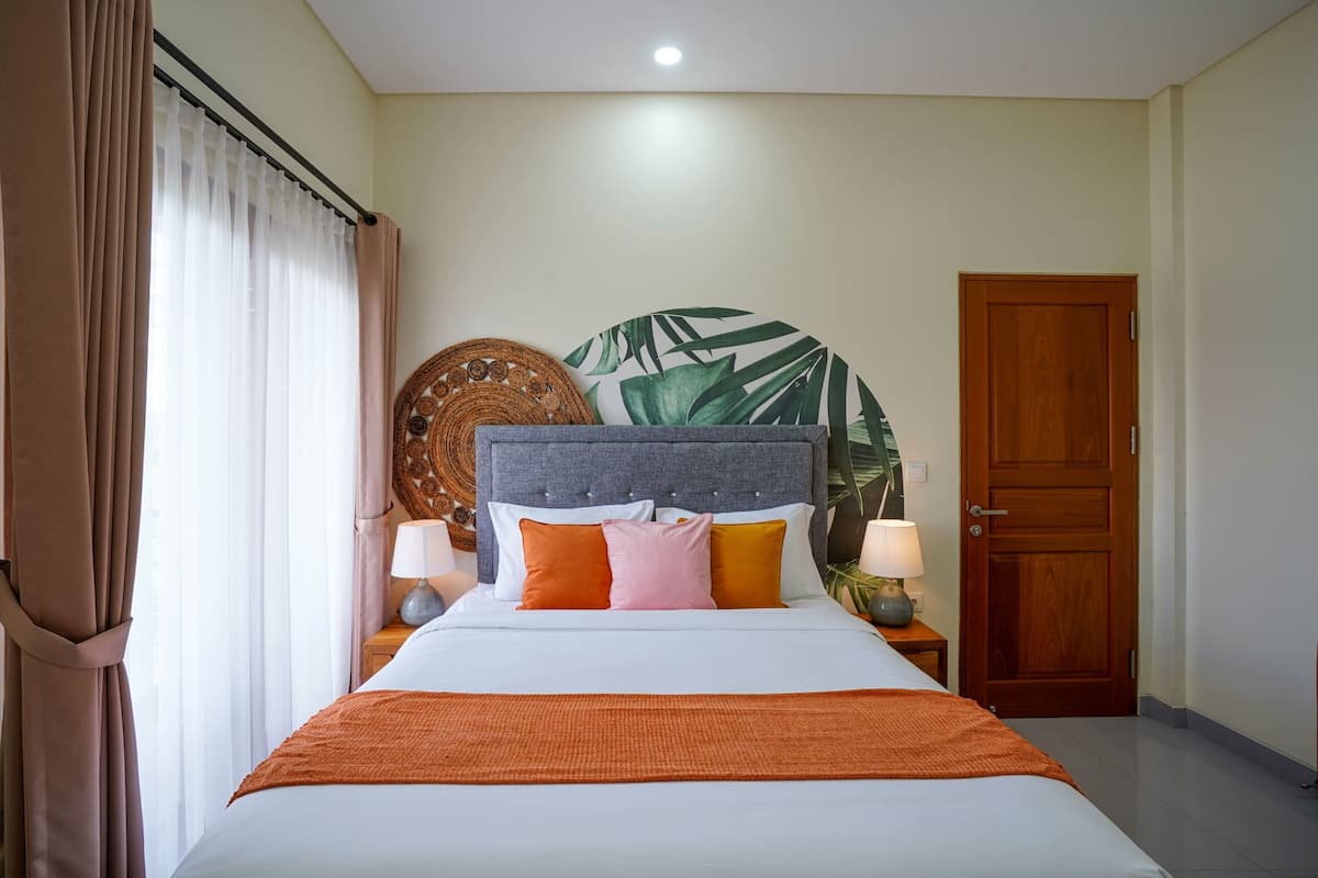 A neatly made bed with a grey headboard, white bedding, and orange accents. There is a large tropical leaf wall art piece above the bed, a wooden door to the right, and a window with sheer curtains on the left.