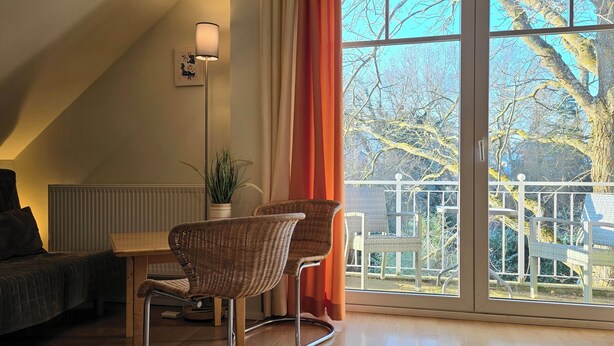 Une pièce avec un canapé de couleur sombre, une petite table, des chaises en osier, un lampadaire et une grande fenêtre donnant sur un jardin et des arbres.