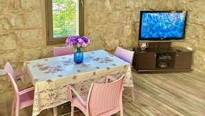 Un comedor con pared de piedra, una mesa con mantel floral, sillas rosas, un jarrón con flores y un televisor sobre un mueble de madera.