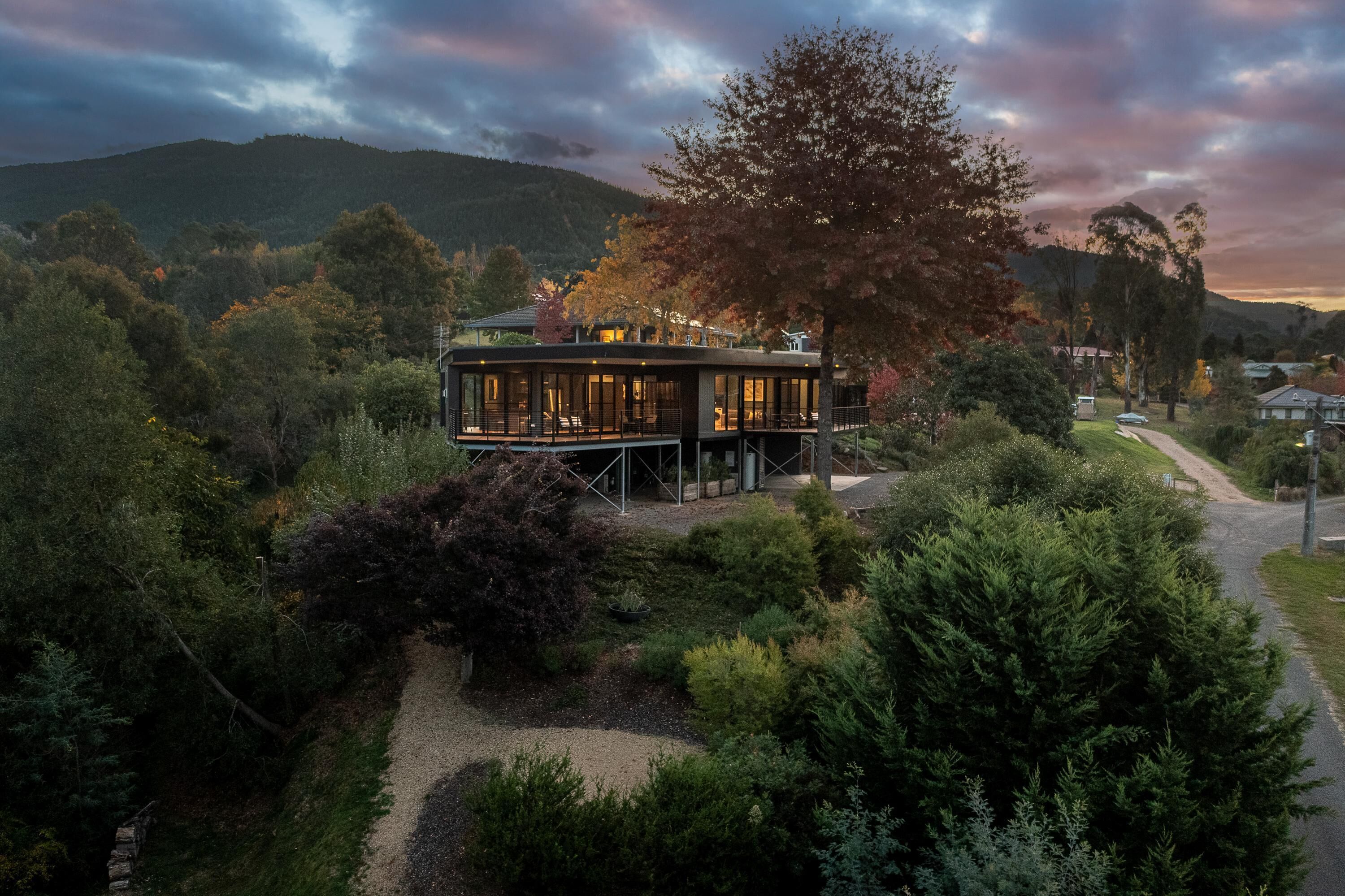 A modern house with a glass facade, surrounded by dense greenery and a mountain backdrop.