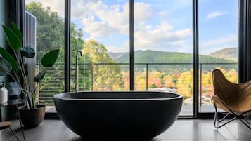 A modern bathroom with a large black bathtub, a wooden chair, and a view of a mountainous landscape through large windows.