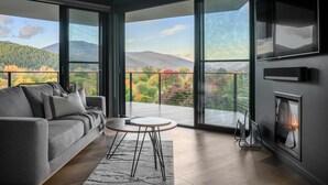 A modern living room with a grey sofa, a round table, and a fireplace. Large windows offer a view of a mountainous landscape. - The View Bright - Apartment 2 - The View Bright has a fabulous outlook and location, being one of the few places in the Bright township with uninterrupted views of the Upper Ovens Valley and Mount Bogong. (Bright)