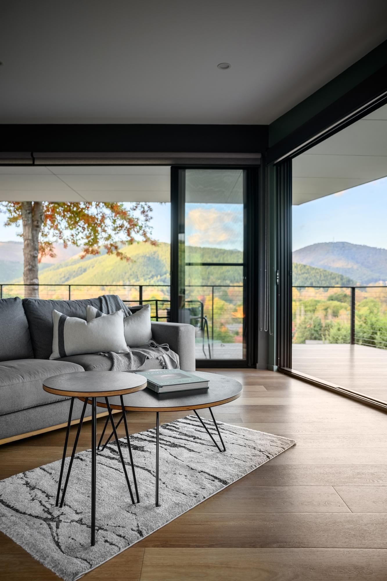 A modern living room with a grey sofa, a round coffee table, and a view of a mountainous landscape through large windows.