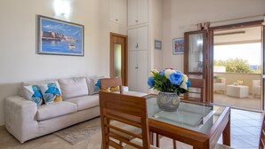 A living room with a sofa, a glass-top table, and a vase with flowers. There is a door leading to an outdoor area with a view. - Sant'Anna house on Brionis estate with large veranda overlooking the sea (Alghero)