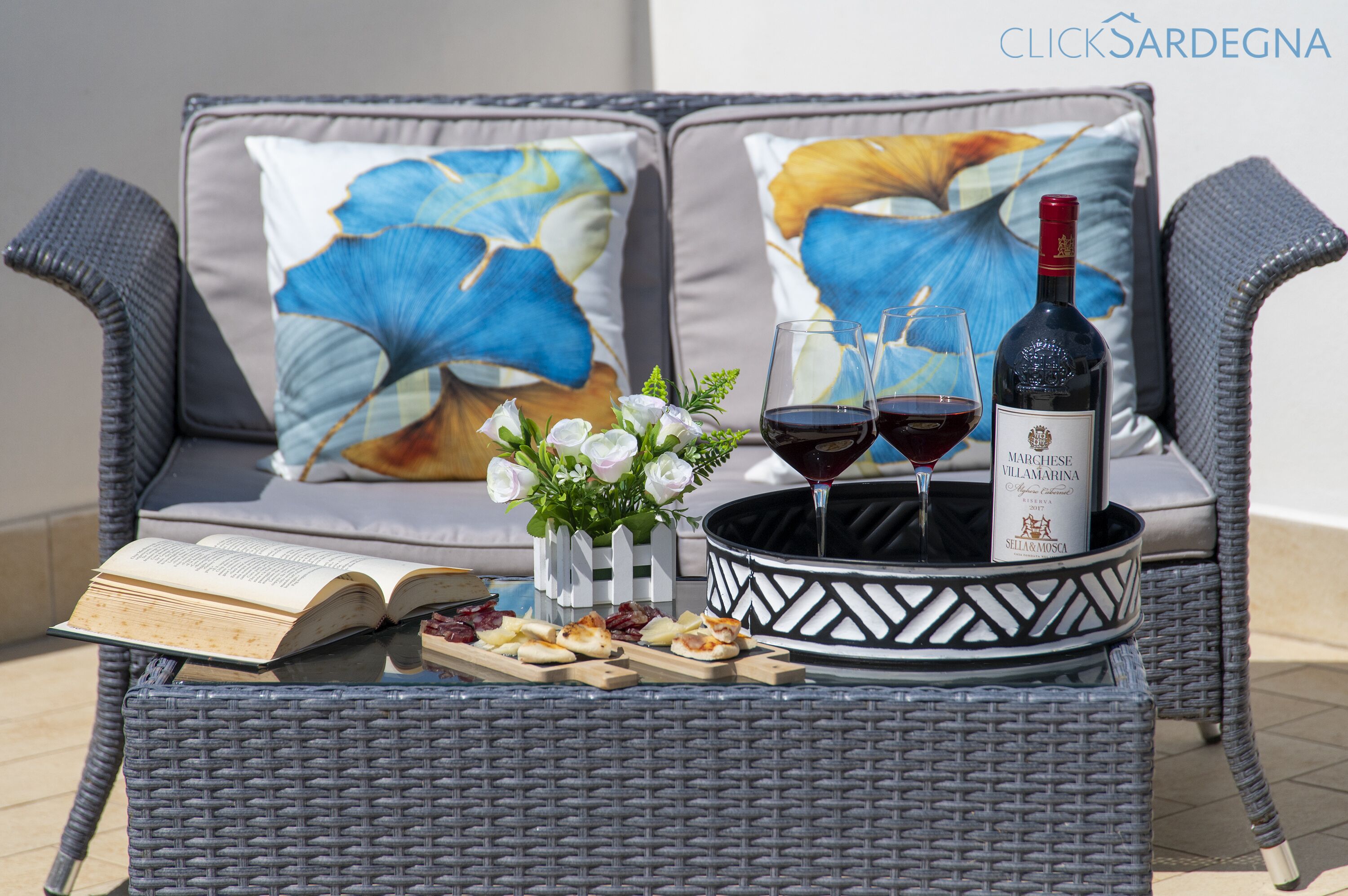 A wicker sofa with cushions, a tray with a bottle of wine, glasses, a book, and a small vase with flowers.