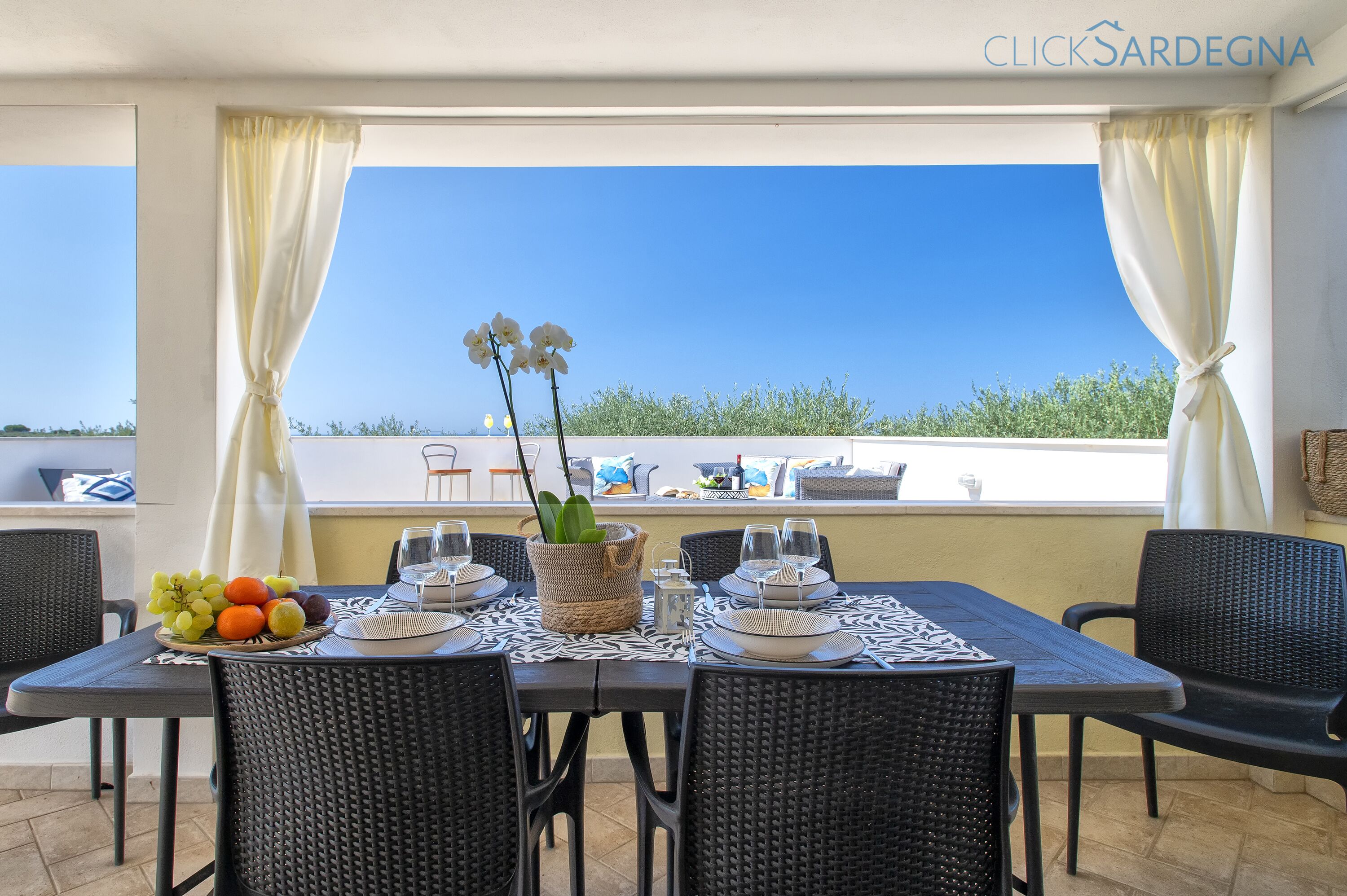 A dining area with a table set for two, featuring a wicker chair, a woven basket with fruit, and a view of the sea.