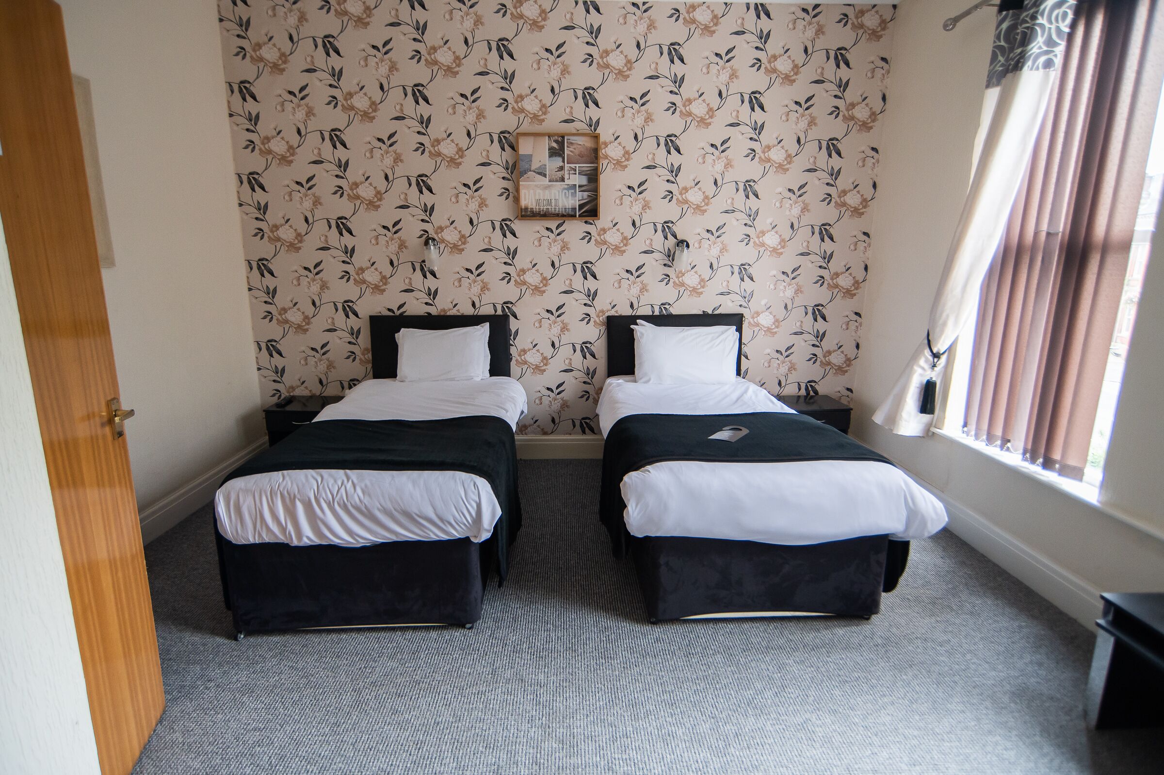 A room with two beds, a window with curtains, a floral wallpaper, and a small picture frame on the wall.