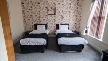 A room with two beds, a window with curtains, a floral wallpaper, and a small picture frame on the wall.