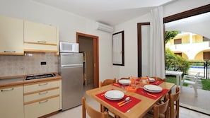A compact kitchen with a refrigerator, microwave, and stove. There is a dining area with a table set for two, and a view of a courtyard with a building in the background. - Cozy Poolside Garden Flat in a Peaceful Residence (San Michele al Tagliamento)
