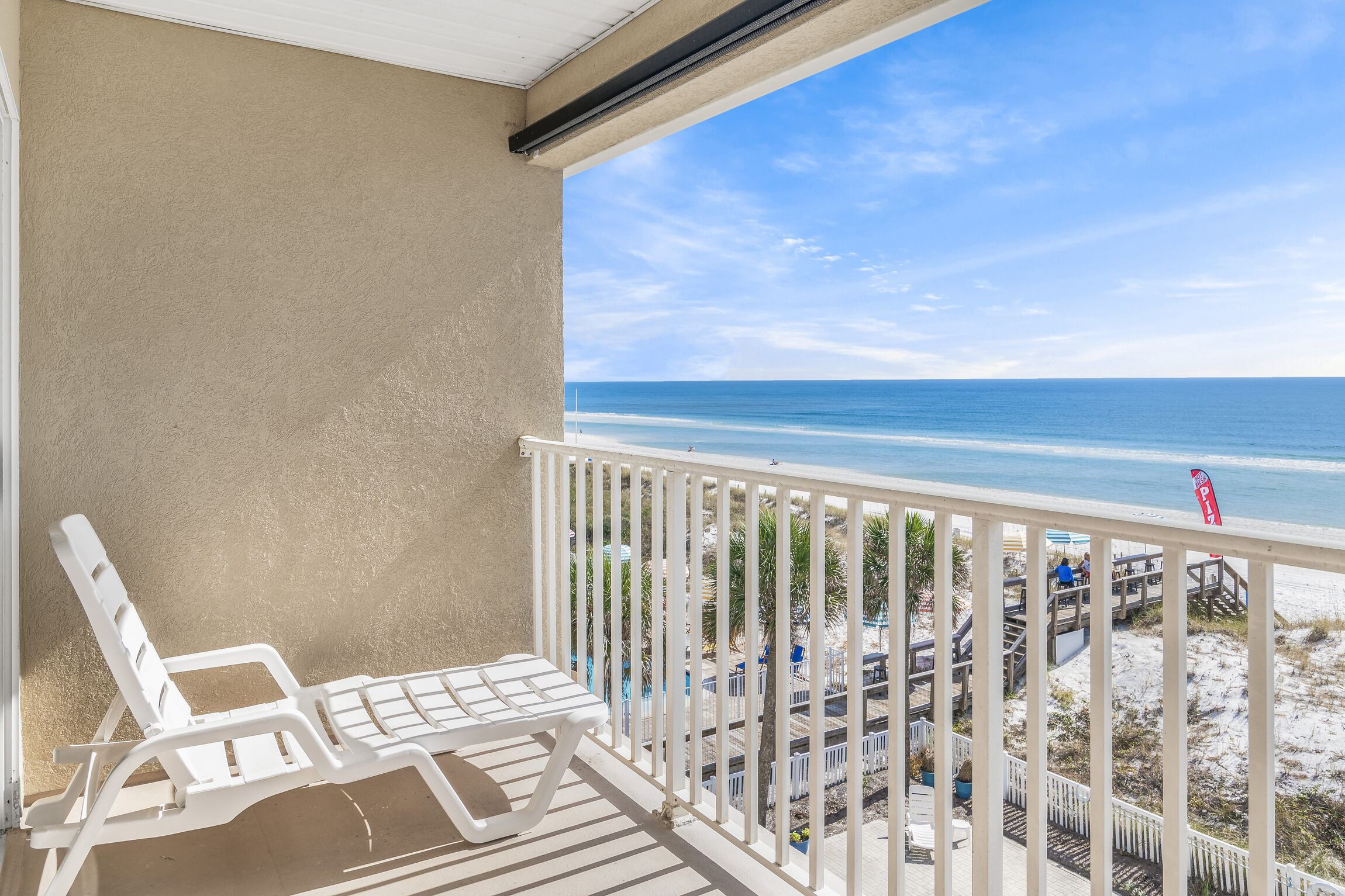 A balcony with a white chair, overlooking a beach with a wooden walkway and beach chairs.