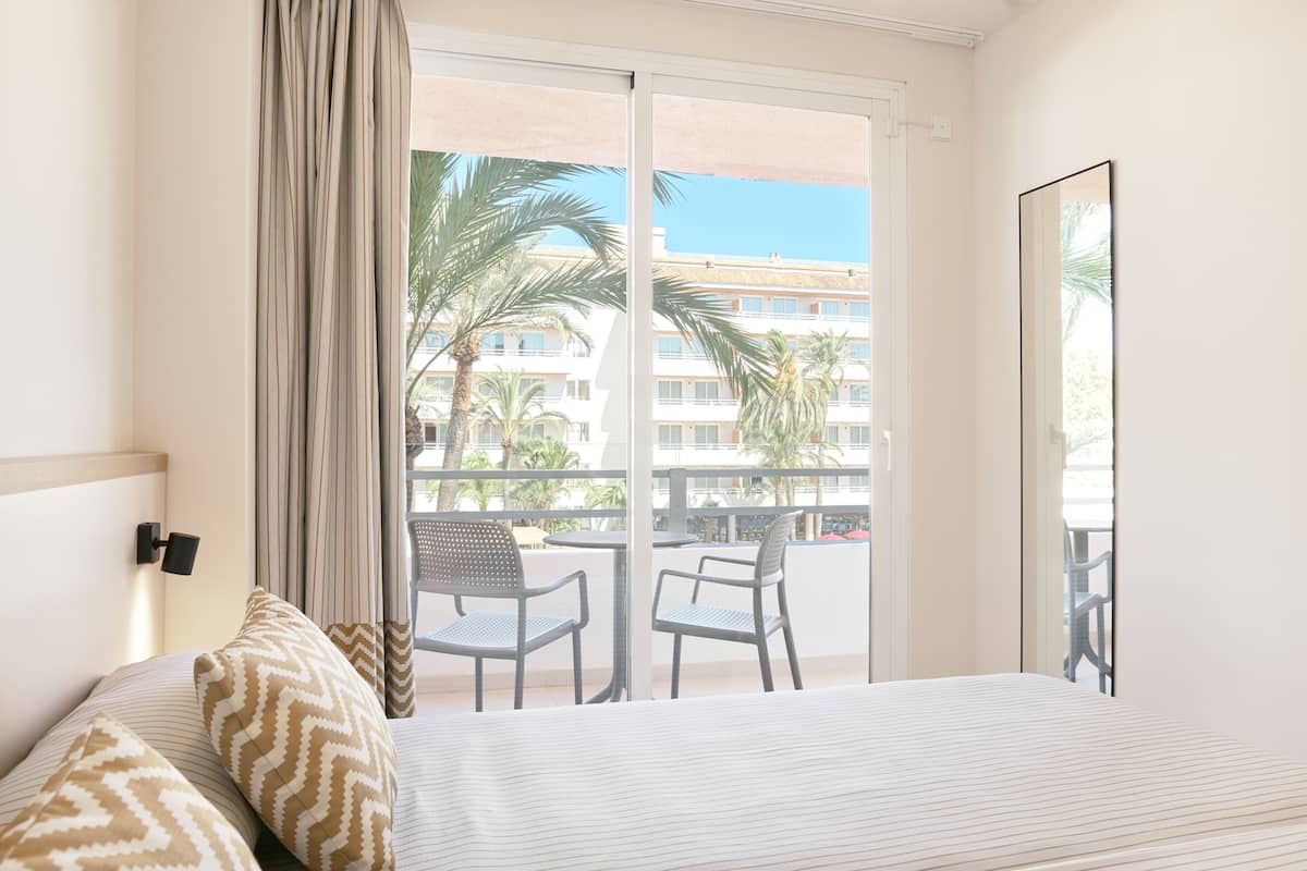 A hotel room with a bed, two chairs, a large window with a view of palm trees and a building, and a mirror on the wall.