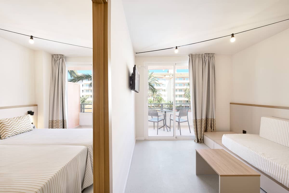 A hotel room with two beds, a bench, a TV, and a balcony with a view of buildings and palm trees.